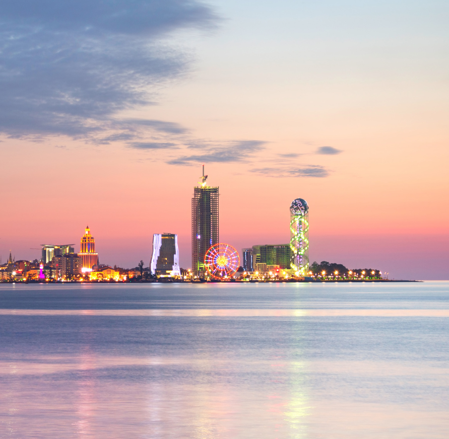 Widok z daleka na bulwar w Batumi w Gruzji o zmierzchu. Budynki są oświetlone. Na pierwszym planie woda.